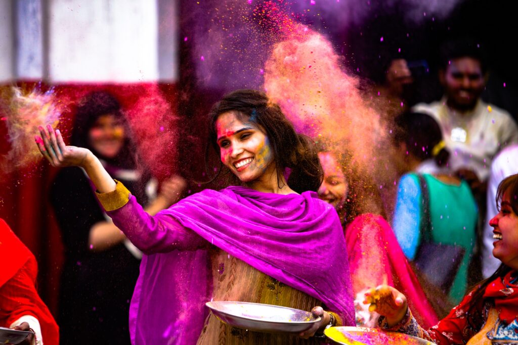 Das Bild zeigt eine dunkelhäutige Frau mit einem lila farbenen Tuch. Sie hält einen Teller und steht vor einer Gruppe anderer Menschen. Das Bild bewirbt den Kurs "Indien - mit allen Sinnen genießen".