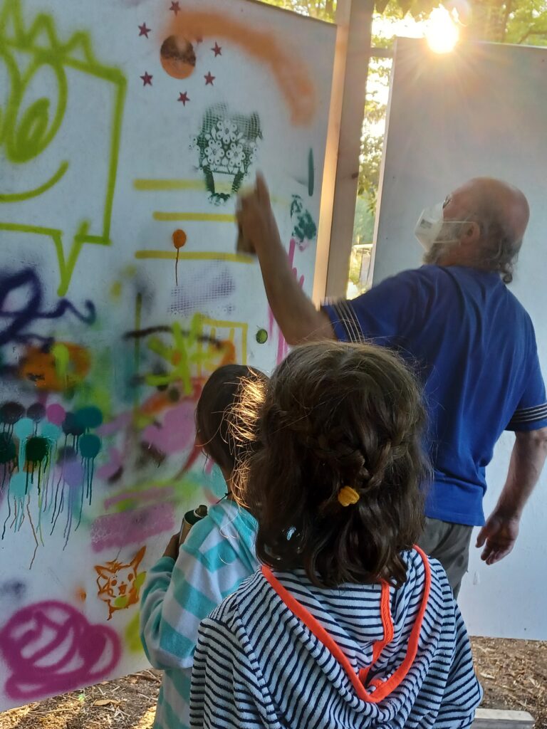 Ein Mann besprüht mit einer Graffiti-Spraydose eine Wand. Davor stehen zwei kleine Mädchen. Alle Personen sind von hinten zu sehen.